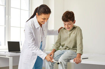 Obraz premium Friendly, smiling doctor examining a happy child. Professional neurologist using a medical hammer to check the knee jerk patellar reflex of a little school boy. Neurology, health checkup concept