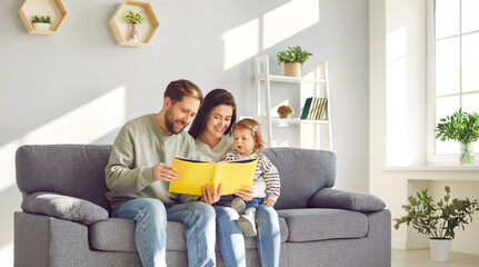 Happy mom and dad reading book to their little daughter. Smiling parents having good time with toddler baby sitting together on sofa at home. Happy parenthood concept