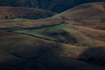 Photo of a scenic view of hills and farm building on the top under sunlight and clouds shadows from the plateau Bermamyt. There are trees in autumn colors on the top of the hills.
