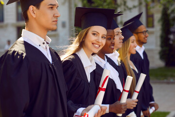 Group of multicultural people in graduation gowns and caps graduate universuty outdoors in campus. They are standing in line. One girl is looking at camera. Graduation from college university concept.