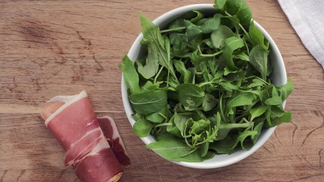 Bol blanco con r&uacute;cula y rulo de jam&oacute;n serrano sobre tabla de madera