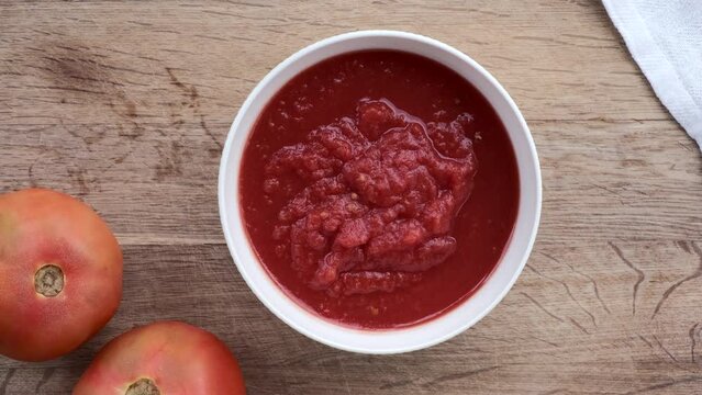 Bol blanco con tomate triturado sobre tabla de madera con dos tomates naturales a su lado.
