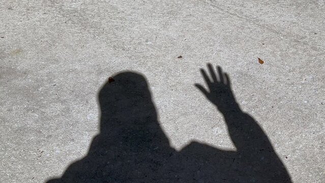 Shadow of man waving hand that is open showing all fingers, standing on concrete