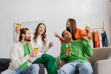 joyful and multicultural group of friends holding glasses of beer while having fun in living room