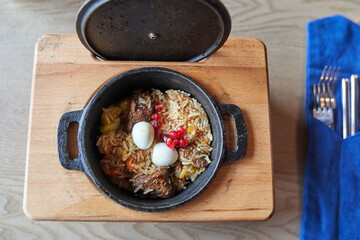 Uzbek national pilaf with meat in a cast-iron frying pan, on a wooden tray with cutlery. Top view with copy space. High quality photo