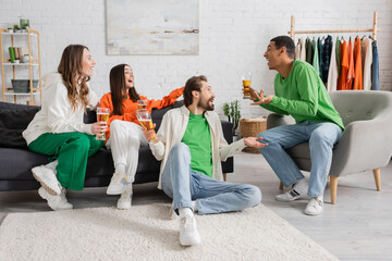Fototapeta premium cheerful multicultural men and women holding glasses of beer while chatting in living room