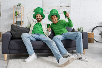 Smiling multiethnic friends in festive hats holding green beer while celebrating saint patrick day
