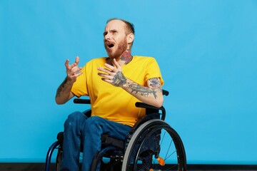 A man in a wheelchair in a T-shirt with tattoos on his arms sits on a blue studio background, a...