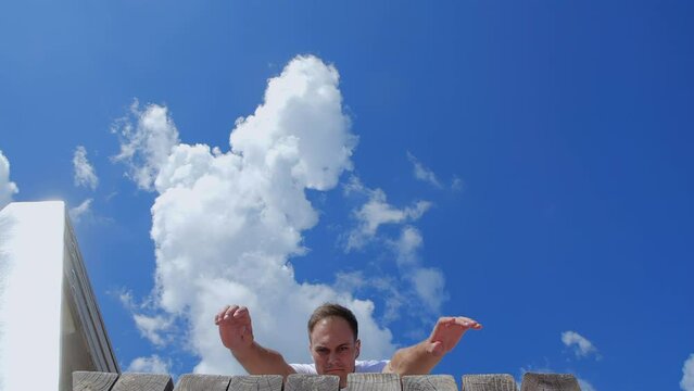 Front view close up  of acrobat performing parkour tricks on benches of glass pedestrian bridge against backdrop of city. Caucasian guy in beige trousers and white T-shirt tumbling. 200 fps slow motio