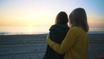 Mother cover his daughter with blanket and hugging on the beach at sunset.