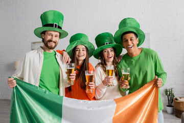 cheerful and multicultural friends in green hats holding glasses of beer and Irish flag while...