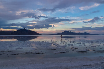 Man sillhoutte walking into sunset of lake Bonneville Salt Flats, Wendover, Western Utah, USA, America. Beautiful summits of Silver Island Mountain range reflecting in water surface, Great Salt Lake