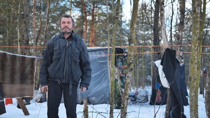 A homeless man poses in the woods in front of his hut.