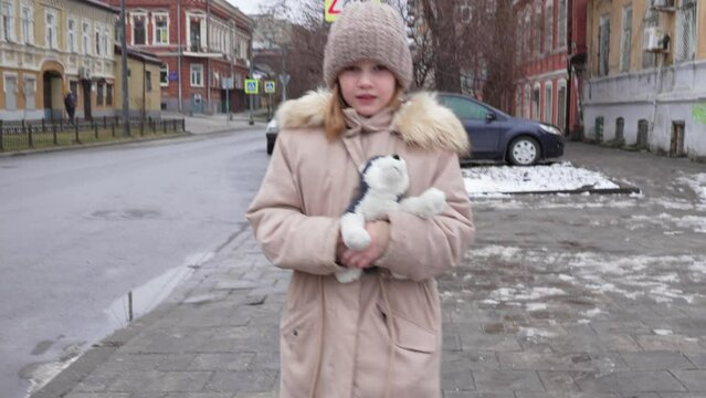 a cute little girl in a hat and coat with a toy walks down a city street.