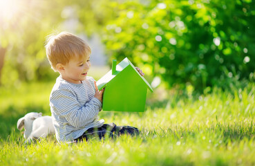 Smiling child sitting on the spring meadow with model of the green house in his hands.