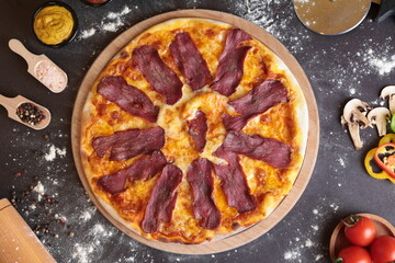 a cheesy pizza with a lot of bacon toppings on it on a table with a pizza cutter and bowl of tomatoes