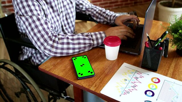 A disabled guy in a wheelchair is working at a laptop at a table, there is a paper cup and a smartphone with a green screen chromakey on the table, documents with a diagram and graphs