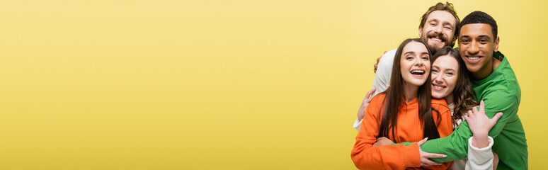 Fototapeta premium Multiethnic friends hugging and smiling at camera isolated on yellow, banner