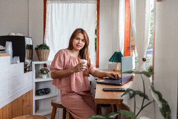 A happy female at a cafe using a laptop in hand and a paper cup of coffee looking at the camera. young white woman with long hair sitting in a coffee shop busy working on her laptop.