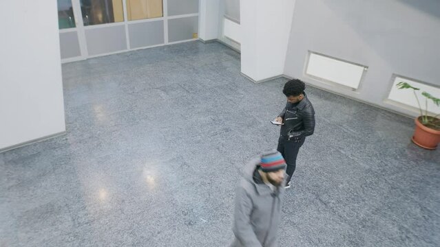 People walking at college lobby. African American man stands and texts by phone. Multi ethnic friends meet. Tracking and monitoring system. Observation and CCTV technologies. Security camera shooting.