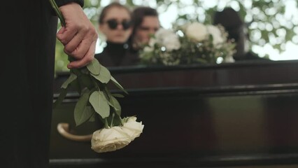 Midsection of unrecognizable woman holding two white roses while standing by wooden coffin at outdoor funeral ceremony