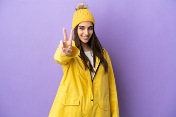 Young caucasian woman wearing a rainproof coat isolated on purple background smiling and showing victory sign