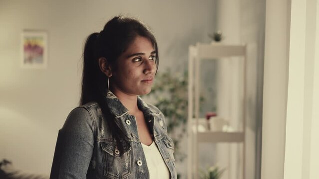 Thoughtful Indian woman with ponytail looks out bright window after waking up in morning. Young housewife in denim jacket thinks about future closeup