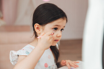 a beautiful little girl in the mirror preening. a little girl is sitting at a children's table and is wearing children's cosmetics.