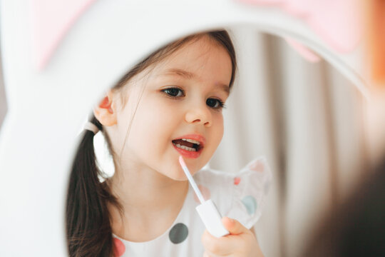 a little girl looks in the mirror and paints her lips with a pink baby glitter.