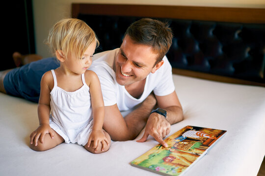 Smiling dad reading book to little daughter pointing finger at pictures