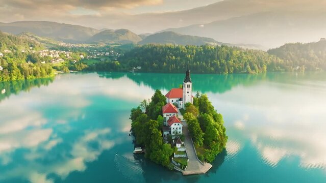 Flying over small island on Bled Lake in Slovenia, warm morning sun, light fog, turquoise water and mountains on horizon. Aerial view of Church of the Assumption of Mary in the center of the lake Bled