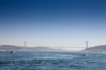 Bosphorus Bridge; also called 15 july martyrs bridge or 15 temmuz sehitler koprusu, seen from afar with boats passing on posporus strait. it's a bridge connecting Asian and European side. .