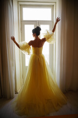 A woman's silhouette in a golden luxurious dress against the background of a window holds a curtain with her hands. Elegant lady in a yellow long silk dress with bare back, back view.