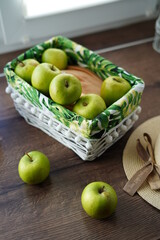 A white wicker basket with green juicy apples on a wooden table near the window. Next to the basket lies a beige hat and a walnut basket. Sunny day. Beautiful background