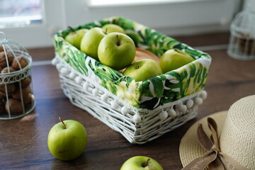 A white wicker basket with green juicy apples on a wooden table near the window. Next to the basket lies a beige hat and a walnut basket. Sunny day. Beautiful background