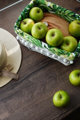 A top view of a white wicker basket with green juicy apples on a wooden table near the window. Next to the basket lies a beige hat and a walnut basket. Sunny day.  Copy space