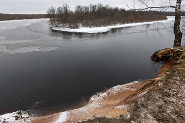 Winter landscape with a partially frozen river. the flow of water. Islet and birch grove. Steep steep bank. Panorama