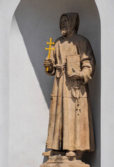 Catholic Monk from Capuchin order with a two-barred cross and Bible book in hands, wear tunic with a big hood on head and a rope as belt. Stone statue of the monk of the Capucins Church in Prague. - 570298845