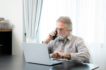 Senior man using mobile phone while sitting at his cozy workplace with laptop at home,Retired male...