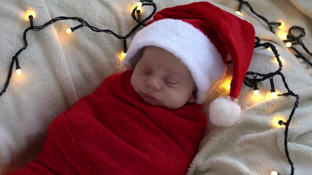 Top View Portrait First Days Of Life Newborn Cute Funny Sleeping Baby In Santa Hat Wrapped In Red Diaper At White Garland Background. Merry Christmas, Happy New Year, Infant, Childhood, Winter Concept