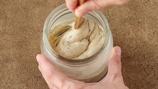 Female hand stirs the wheat sourdough starter with a wooden spoon in a glass jar. Natural yeast for making rye or wheat artisan bread. Fermentation product. Close up. 
