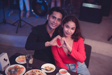 A loving Asian couple forms a heart with their hands as they pose for a picture while at dinner. Boyfriend and girlfriend eating out and making memories on valentine's day.