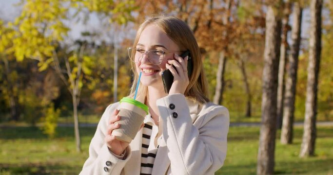 Portrait of a beautiful girl in glasses talking on the phone, laughing and drinking a drink from a large glass, walking in the park in the evening