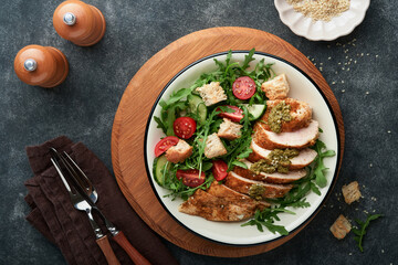 Fresh salad with grilled chicken fillet, lettuce, arugula, tomatoes, cucumber and pesto sauce, sesame seeds with olive oil in white bowl on black slate background. Healthy lunch menu. Top view.