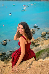 Fototapeta na wymiar A girl sits on a cliff.A woman turns to the camera.A portrait near the sea.A high cliff.A lady in red.The wind blows on her hair.A beautiful azure sea.Emotion of joy and happiness.Rest on the sea.