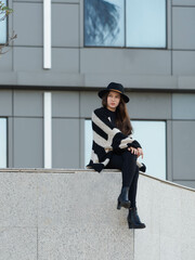 Stylish young girl with hat is sitting on overhanging wall with legs crossed. Modern building in background. Portrait.