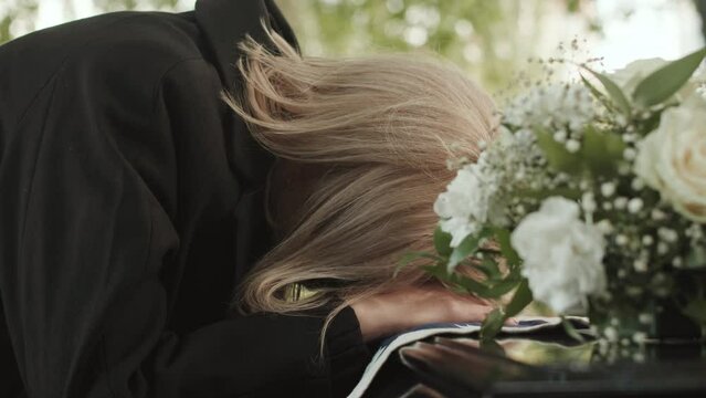 Grieving widow in black coat and eyeglasses putting her head on casket lid with US burial flag on it, saying goodbye to her beloved husband during outdoor funeral ceremony