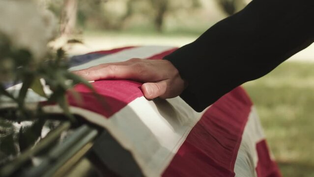 Closeup of unrecognizable male hand on casket lid with unfolded US burial flag on it at outdoor funeral ceremony