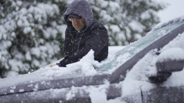 Man cleaning snow-covered car with brush during heavy snowfall. Snowstorm, bad winter weather, natural disaster. Snowstorm in winter season. Vehicle care in blizzard