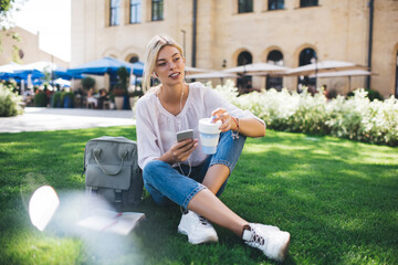 Charming Caucasian woman 20s in headphones listening audio podcast in summer park, millennial female blogger with smartphone device enjoying music playlist during coffee break at green grass